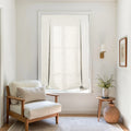 A bright, minimalist interior corner with a cream upholstered wooden armchair, a white fabric Roman blind on the window, a wooden side table with a plant and woven basket, and a brass wall sconce