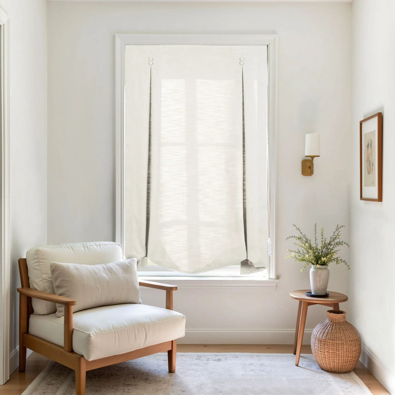 A bright, minimalist interior corner with a cream upholstered wooden armchair, a white fabric Roman blind on the window, a wooden side table with a plant and woven basket, and a brass wall sconce