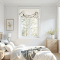 Bright photograph of a minimalist bedroom featuring a bed with white linen and a gray throw, natural wood furniture including a nightstand and dresser, a window with a gray Roman shade overlooking trees, a framed botanical print, and a vase of dried yellow flowers