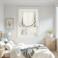 A bright bedroom interior showcasing a large window with a white linen balloon roman shade, a bed with white bedding and a soft grey throw, light wood furniture, and a framed yellow botanical print