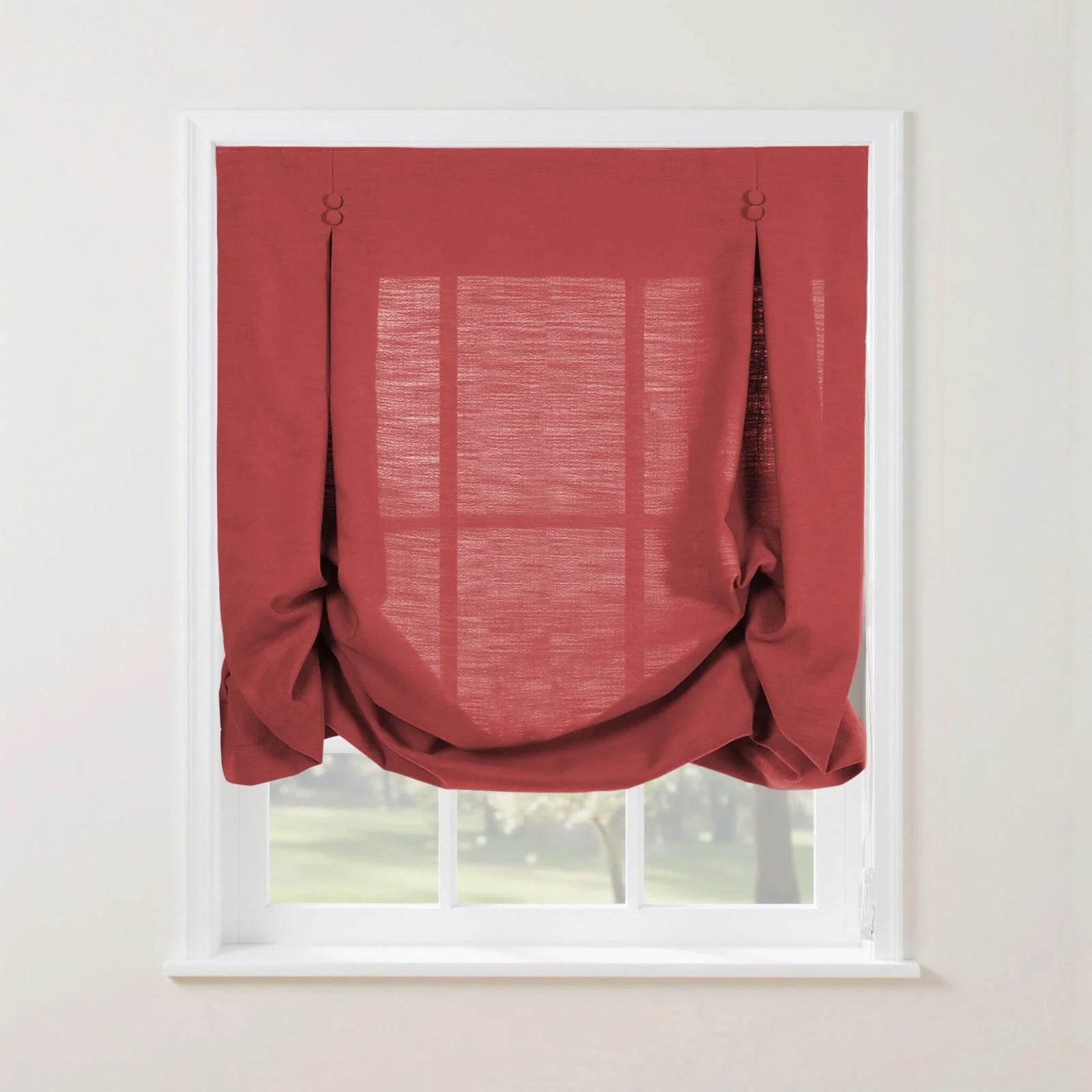 Red linen Roman blind with decorative button and loop pleats and a softly gathered bottom on a white window frame revealing a blurred green outdoor scene