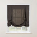Photograph of a dark grey linen Roman blind featuring decorative button loops at the top and a voluminous gathered bottom swag, set against a white window frame with a blurred green garden visible through its semi-sheer fabric