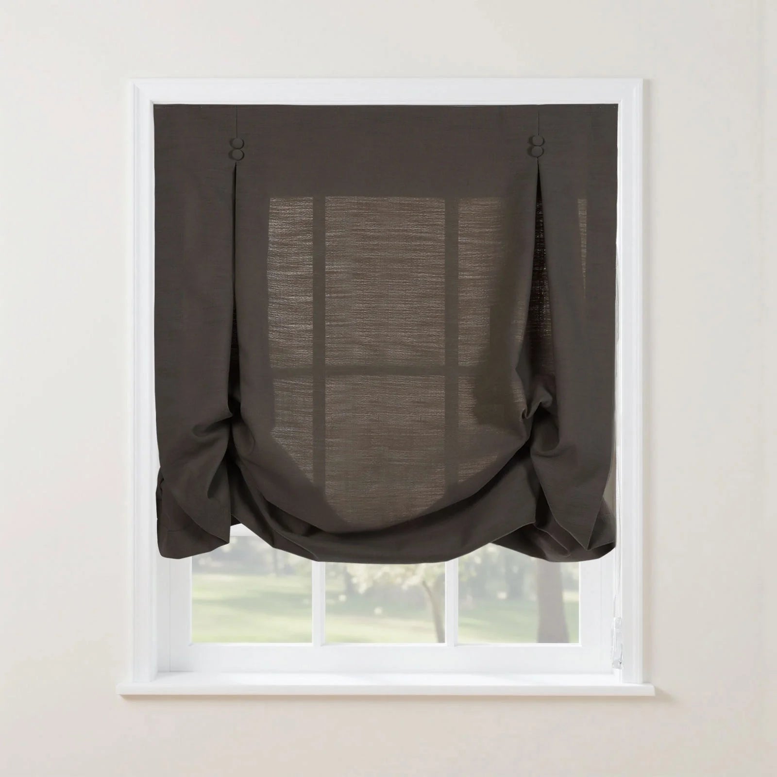 Photograph of a dark grey linen Roman blind featuring decorative button loops at the top and a voluminous gathered bottom swag, set against a white window frame with a blurred green garden visible through its semi-sheer fabric