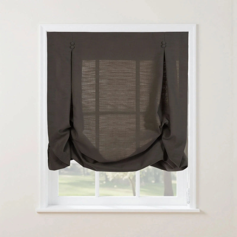 Photograph of a dark grey linen Roman blind featuring decorative button loops at the top and a voluminous gathered bottom swag, set against a white window frame with a blurred green garden visible through its semi-sheer fabric