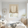 A bright minimalist bedroom featuring a window with a beige relaxed Roman shade, white bedding and grey throw, light wood furniture, a framed botanical print with yellow flowers, and a white ribbed ceramic lamp and vase