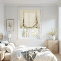 Photography of a bright minimalist bedroom interior showcasing a window with a beige linen balloon roman shade light wood furniture white bedding with a grey throw and a framed yellow botanical print