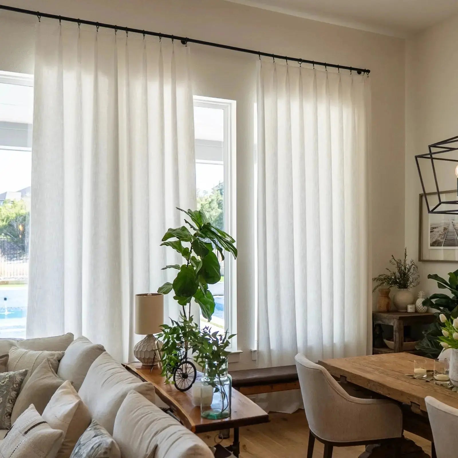 A bright modern living and dining room with floor-to-ceiling white sheer curtains, a prominent fiddle leaf fig plant, a natural wood console table, and a backyard pool visible through the window