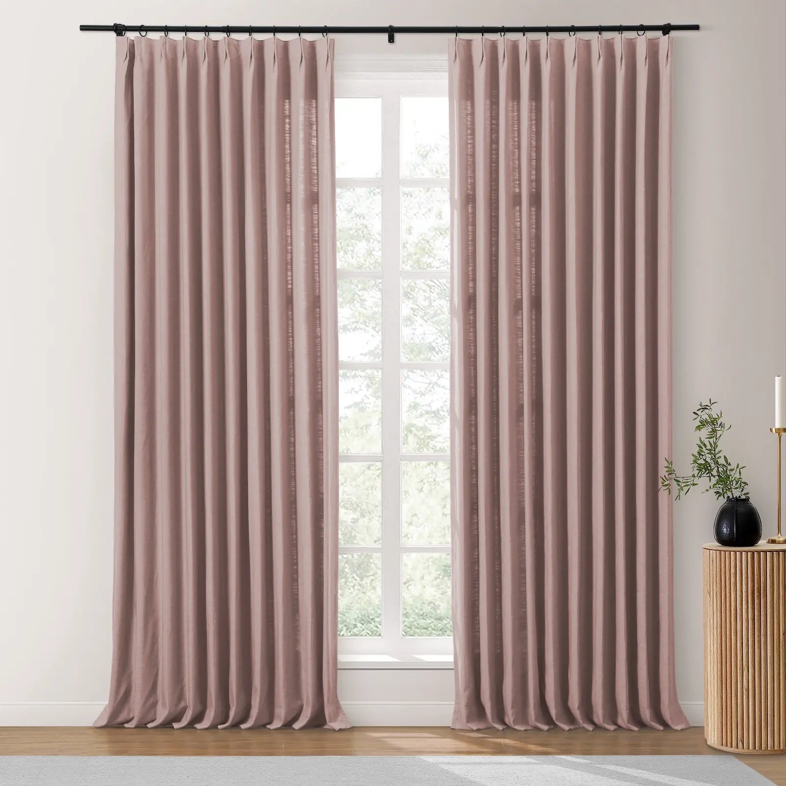Dusty rose linen-look pleated curtains on a black rod frame a window with a lush green outdoor view, complemented by a fluted wooden side table holding a black vase with greenery and a gold candle holder