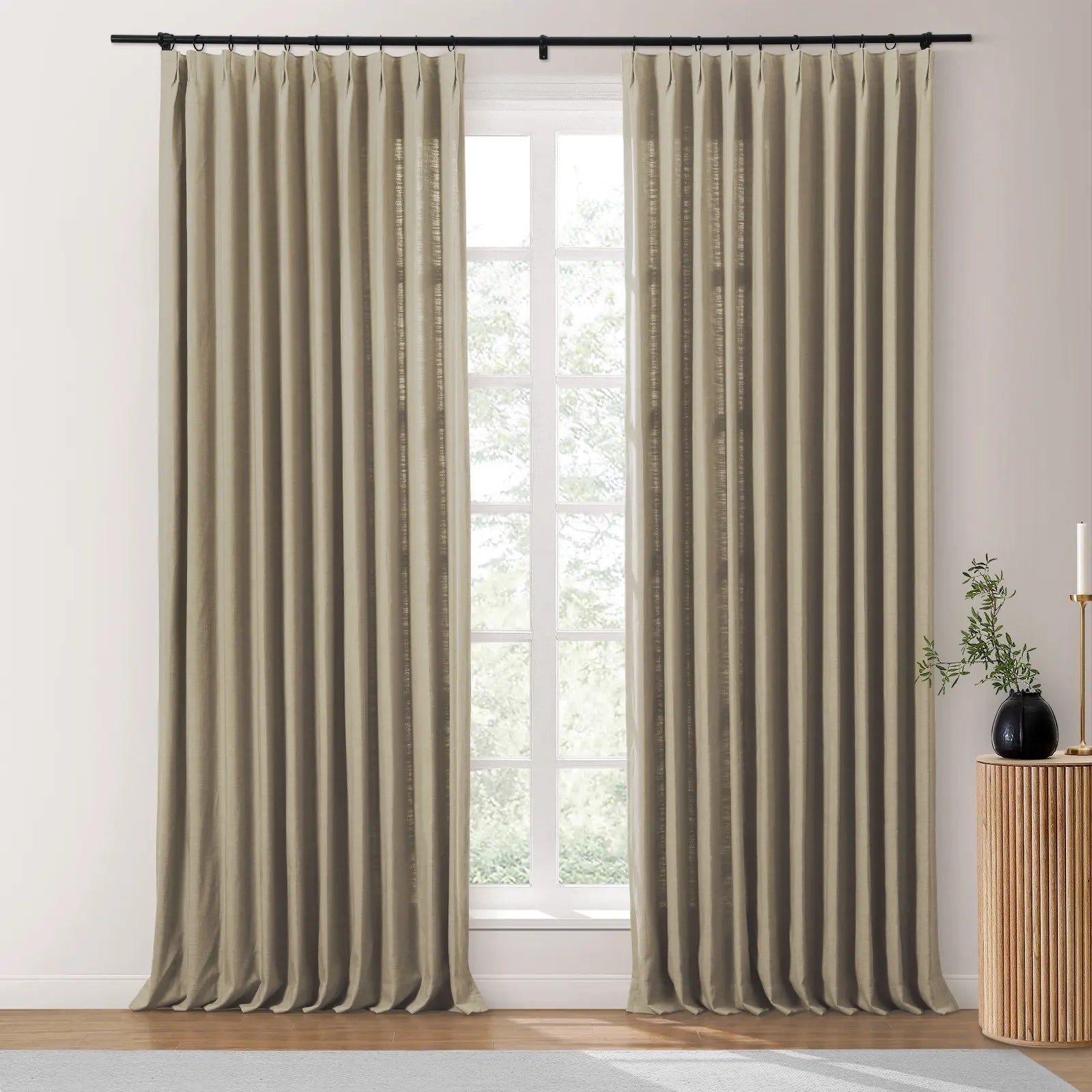 Light beige pinch pleat linen curtains hang on a black rod framing a large white window revealing green foliage beyond with a fluted wood side table, black vase, and gold candlestick to the right