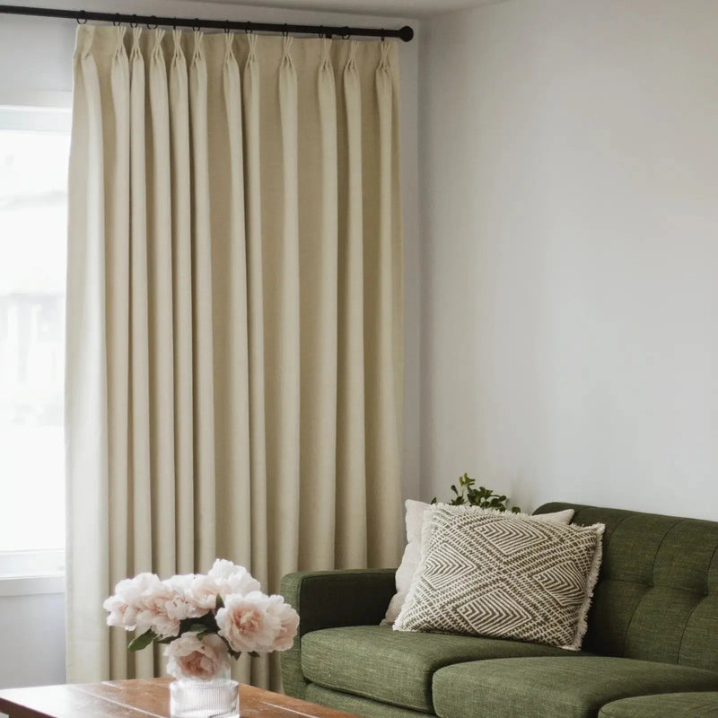 Photograph of a cozy living room interior featuring an olive green tufted sofa with a patterned cushion, elegant beige pinch-pleat drapes, and a vase of light pink peonies on a wooden coffee table