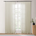 Bright room featuring off-white sheer pleated curtains on a black rod over a window revealing green trees, with a fluted wood side table holding a black vase of green foliage and a brass candlestick