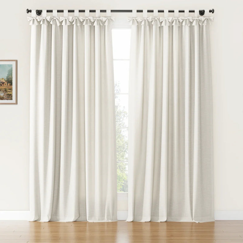 Off-white linen-look tie-top curtains featuring decorative bows on a black rod in a bright room with a warm wooden floor and a glimpse of a small framed landscape painting