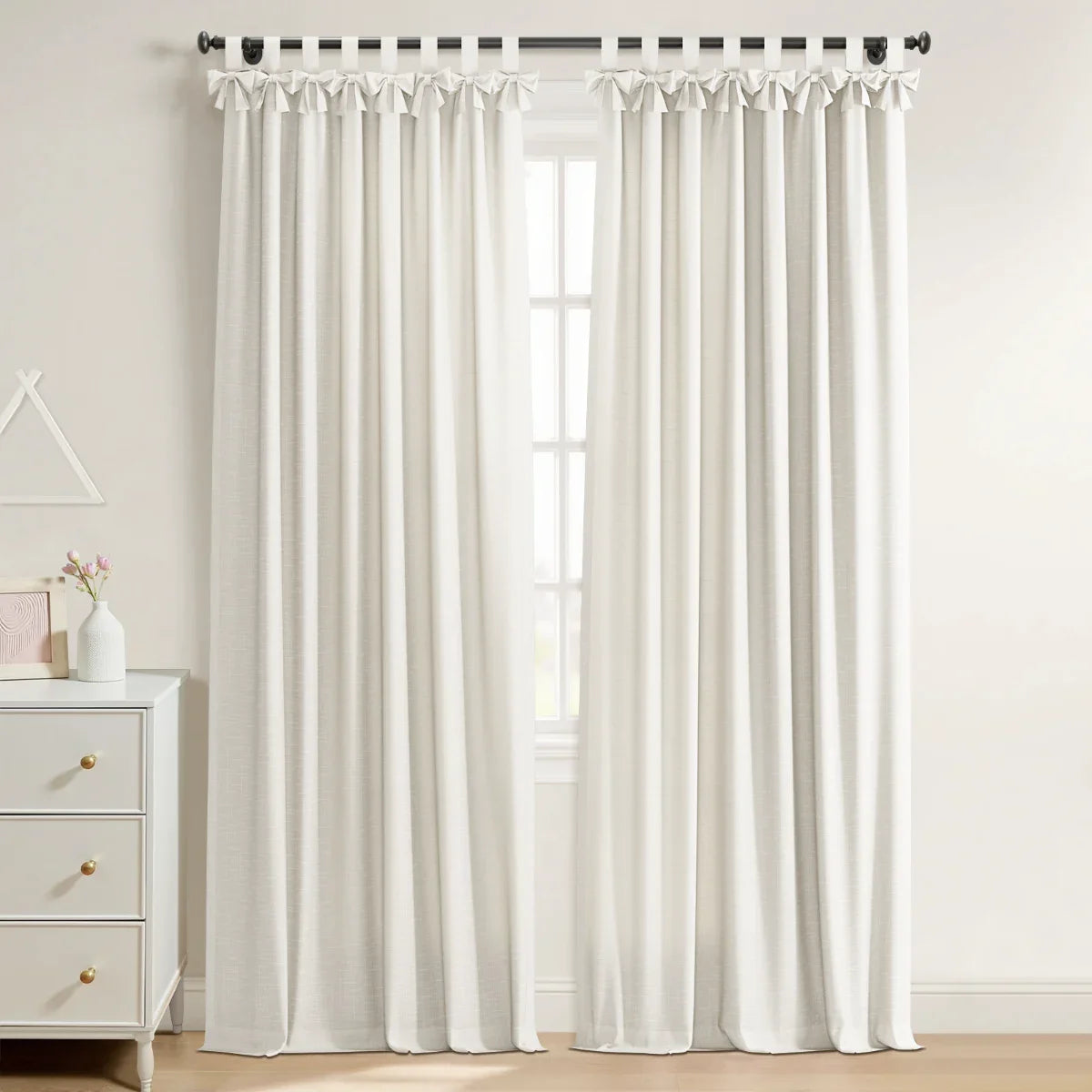 White linen-blend tie-top curtains on a black rod in a bright room, next to a white three-drawer dresser with brass pulls and pink flowers