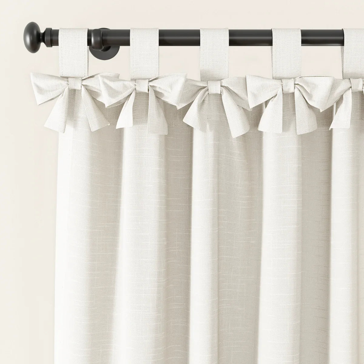 A cream linen-textured curtain with decorative bow-tie top tabs hangs from a black curtain rod with a round finial