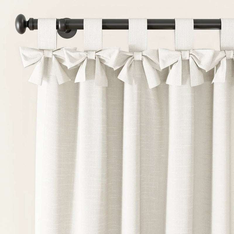 A cream linen-textured curtain with decorative bow-tie top tabs hangs from a black curtain rod with a round finial
