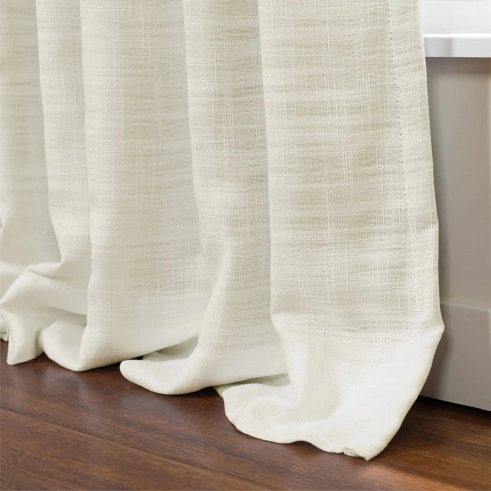 Photograph of a white textured sheer curtain with heavy folds pooling on a dark brown wooden floor next to a white window sill