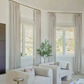 Photograph of a modern living room interior featuring two curved cream armchairs with patterned throw pillows, a faux olive tree, and beige floor-to-ceiling drapes, overlooking a dry landscape with a white fence