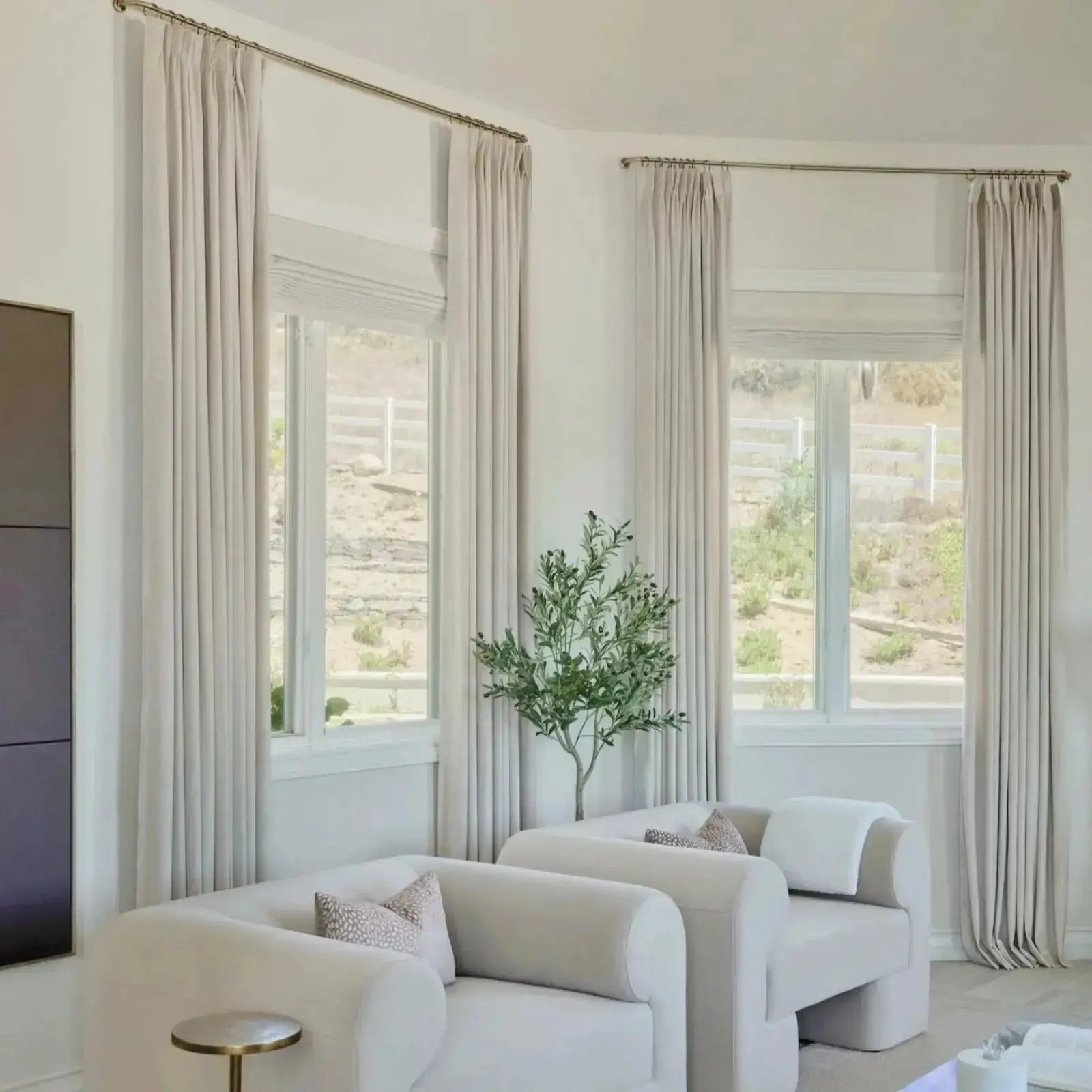 Photograph of a modern living room interior featuring two curved cream armchairs with patterned throw pillows, a faux olive tree, and beige floor-to-ceiling drapes, overlooking a dry landscape with a white fence