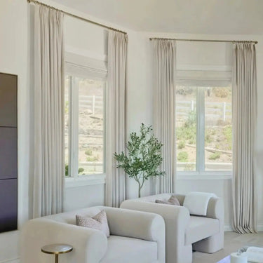 Photograph of a modern living room interior featuring two curved cream armchairs with patterned throw pillows, a faux olive tree, and beige floor-to-ceiling drapes, overlooking a dry landscape with a white fence