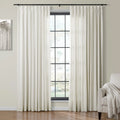 Full-length off-white pleated curtains on a black rod frame a grid window with an outdoor view, flanked by a minimalist tree painting and a beige armchair with a textured throw