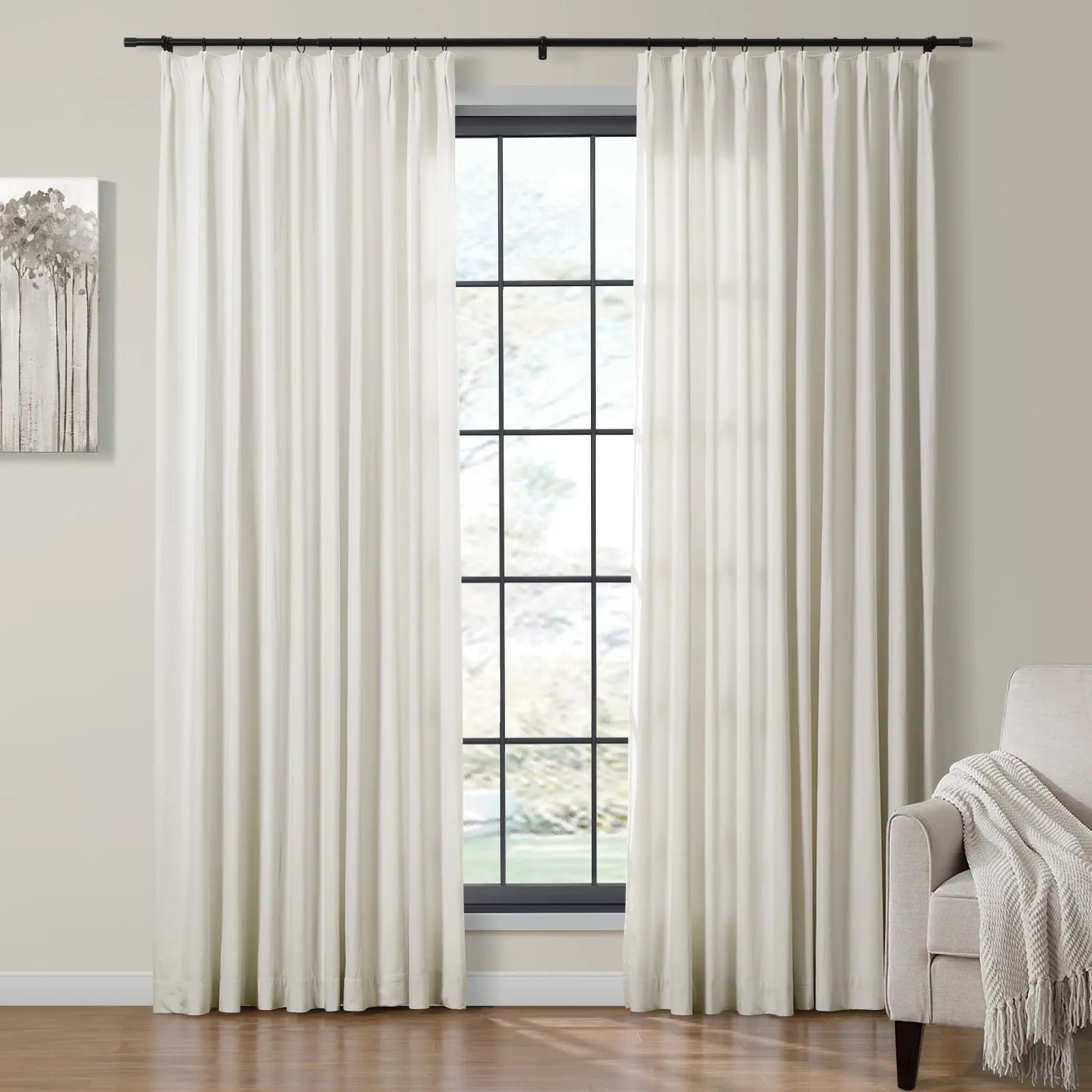 Full-length off-white pleated curtains on a black rod frame a grid window with an outdoor view, flanked by a minimalist tree painting and a beige armchair with a textured throw