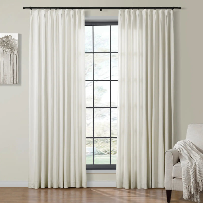 Full-length off-white pleated curtains on a black rod frame a grid window with an outdoor view, flanked by a minimalist tree painting and a beige armchair with a textured throw