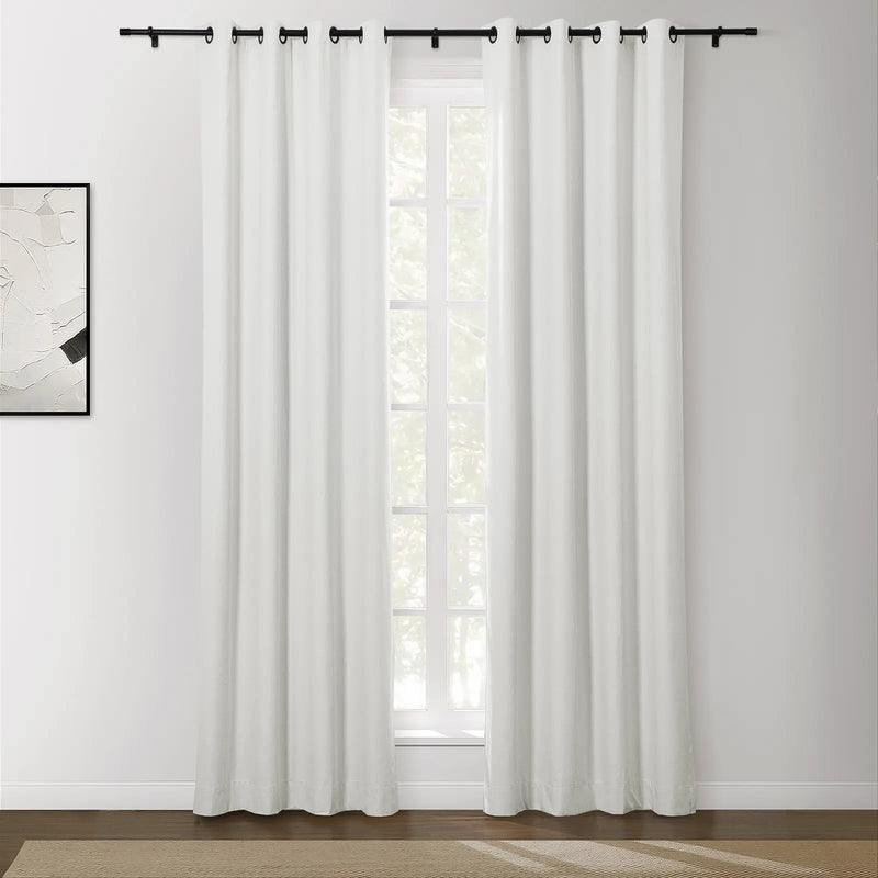 Photograph of a modern interior with a window dressed in white blackout curtains featuring black grommets and rod, an abstract white and black framed artwork on the left wall, and a light brown textured rug on a dark wood floor
