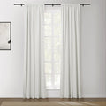 A bright room featuring white floor-length curtains on a black rod over a window looking out to green foliage, alongside a framed abstract artwork and a beige textured rug on a dark wood floor