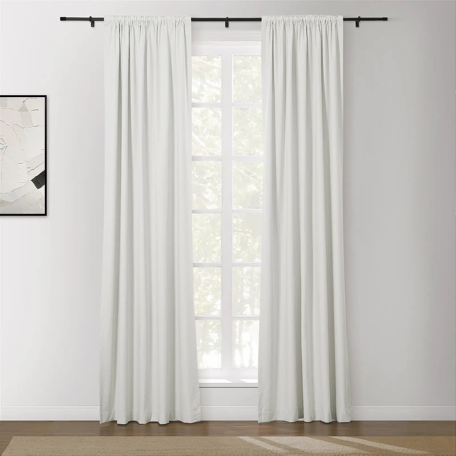 A bright room featuring white floor-length curtains on a black rod over a window looking out to green foliage, alongside a framed abstract artwork and a beige textured rug on a dark wood floor