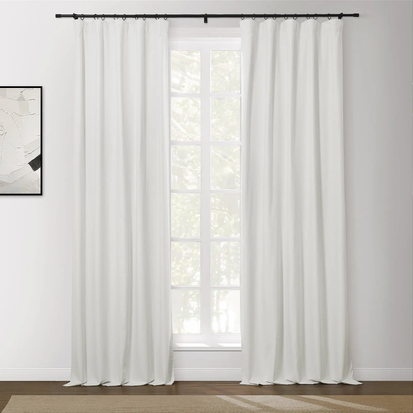 Photograph of a brightly lit interior window featuring white blackout curtains on a black rod, an abstract white and black artwork on the left wall, and a patterned brown rug on a hardwood floor