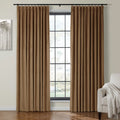 An inviting room showcasing two brown pleated curtains on a black rod framing a multi-pane window with blurred outdoor greenery, an abstract grey tree artwork on the left, and a beige armchair with a white fringed throw blanket on the right, all on a gleaming hardwood floor
