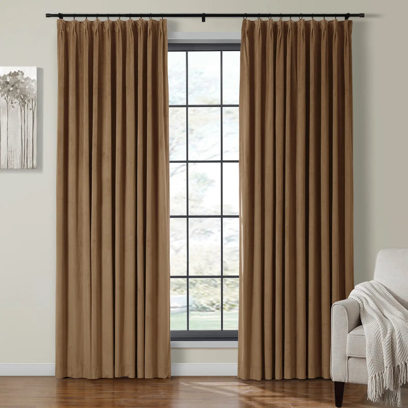An inviting room showcasing two brown pleated curtains on a black rod framing a multi-pane window with blurred outdoor greenery, an abstract grey tree artwork on the left, and a beige armchair with a white fringed throw blanket on the right, all on a gleaming hardwood floor