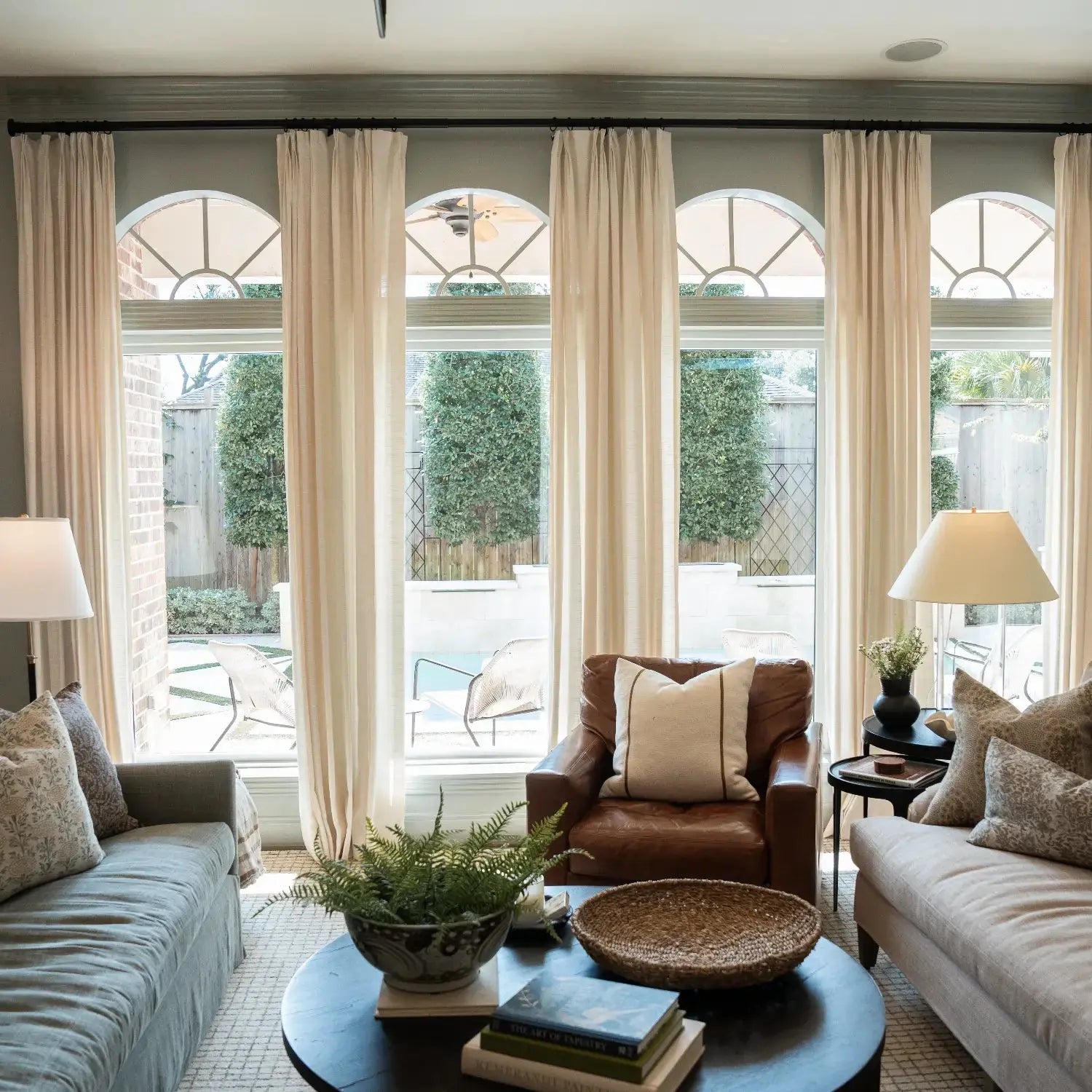 Photograph of a cozy living room featuring multiple tall arched windows overlooking a backyard pool, furnished with a brown leather armchair, two light-toned sofas, and a dark circular coffee table adorned with a fern and books