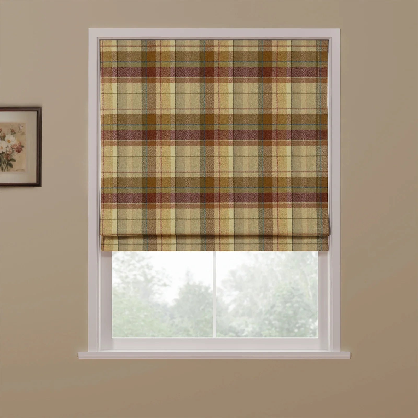 A Roman blind featuring a multi-toned beige, brown, and maroon plaid pattern with subtle blue lines partially covers a window overlooking blurred trees, with a small framed floral print on the adjacent beige wall