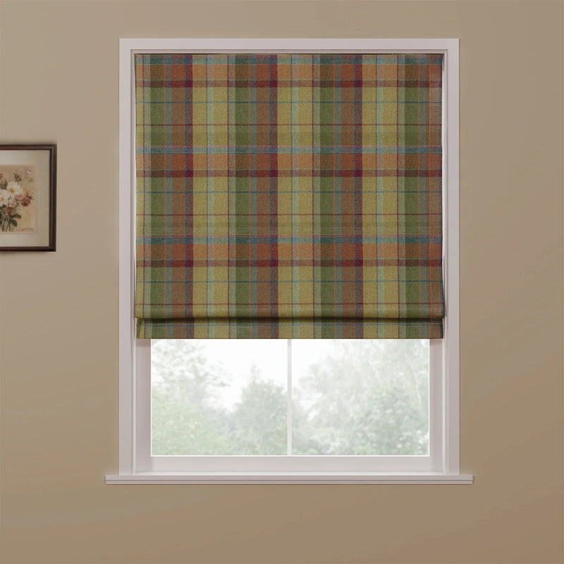 A Roman blind with an earthy plaid pattern in muted greens, browns, rust, and blue covering a window, showing blurred trees outside and a small framed floral picture on a beige wall
