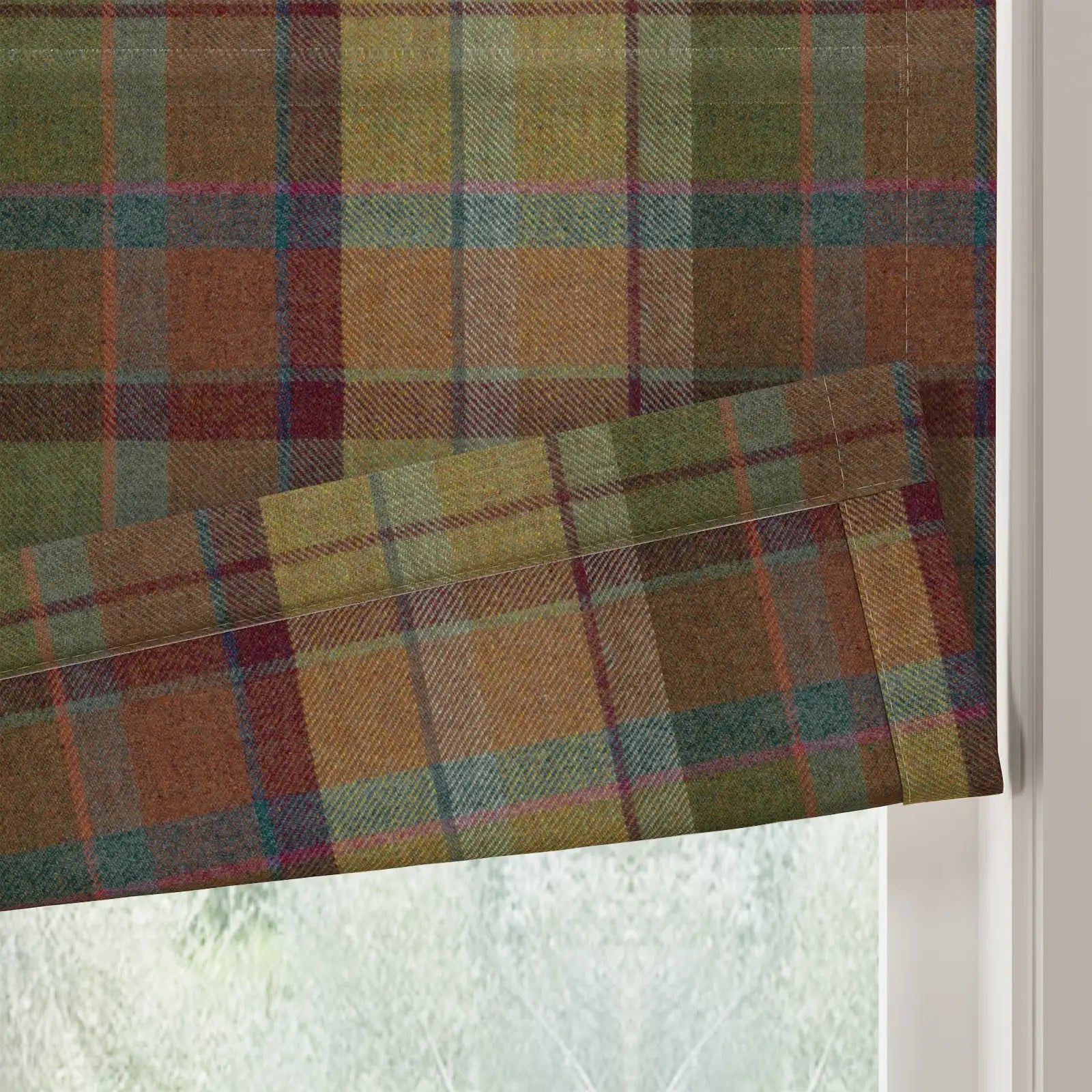 Textile Roman blind in an earthy plaid pattern with olive green, rust brown, deep teal, and fine magenta stripes, partially lowered in a window overlooking a blurred outdoor scene