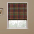 A partially lowered Roman blind featuring a multi-color tartan plaid pattern with earthy reds, greens, and browns, set in a white window frame against a beige wall with a floral art print, overlooking a blurred green outdoor scene