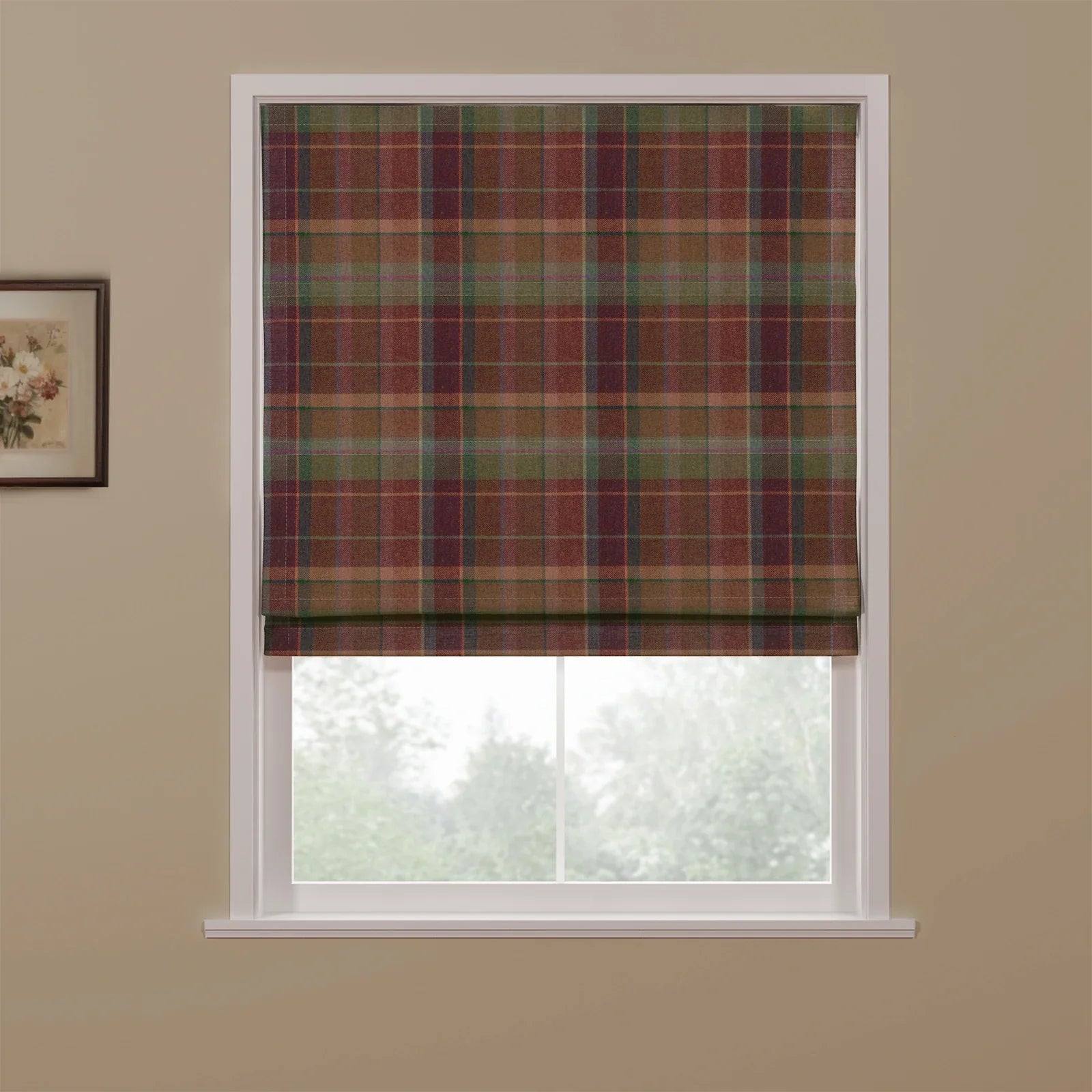 A partially lowered Roman blind featuring a multi-color tartan plaid pattern with earthy reds, greens, and browns, set in a white window frame against a beige wall with a floral art print, overlooking a blurred green outdoor scene