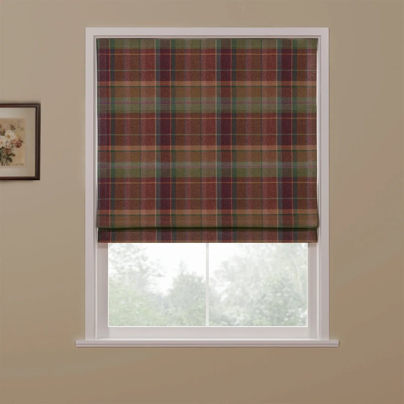 A partially lowered Roman blind featuring a multi-color tartan plaid pattern with earthy reds, greens, and browns, set in a white window frame against a beige wall with a floral art print, overlooking a blurred green outdoor scene