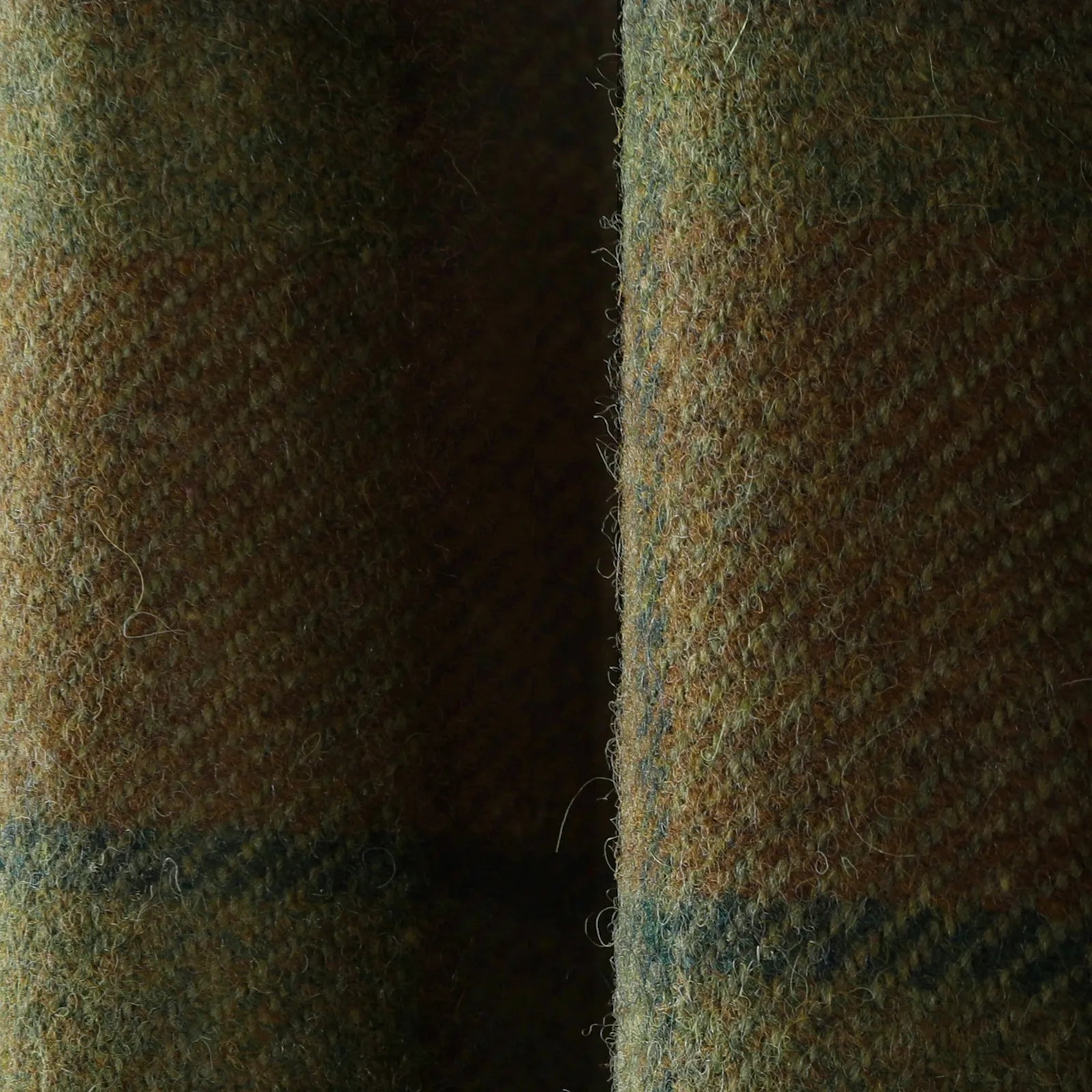 Photograph of a close-up textured wool tweed fabric in earthy olive green, brown, and hints of dark blue, featuring a prominent vertical fold and visible fuzzy surface fibers