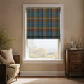 A room interior features a window with a teal, mustard yellow, and plum tartan Roman blind, offering a view of a hazy autumn landscape, flanked by a wooden daybed with rattan details and a dark wood side table adorned with brass candlesticks and a white vase