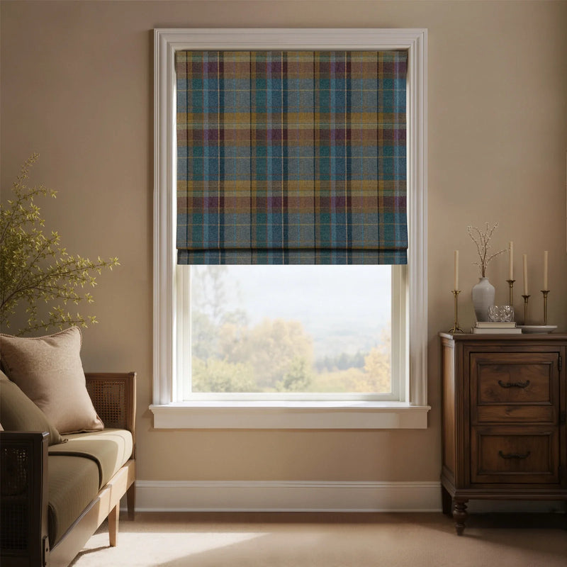 A room interior features a window with a teal, mustard yellow, and plum tartan Roman blind, offering a view of a hazy autumn landscape, flanked by a wooden daybed with rattan details and a dark wood side table adorned with brass candlesticks and a white vase
