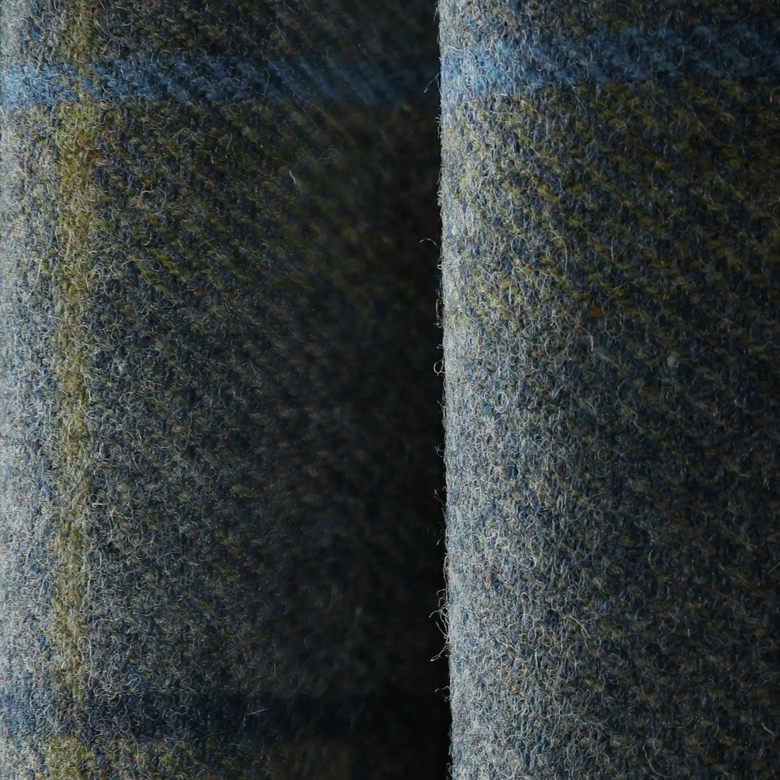 A close-up of folded dark grey-blue wool fabric with visible textures, featuring subtle olive green and light blue threads woven throughout