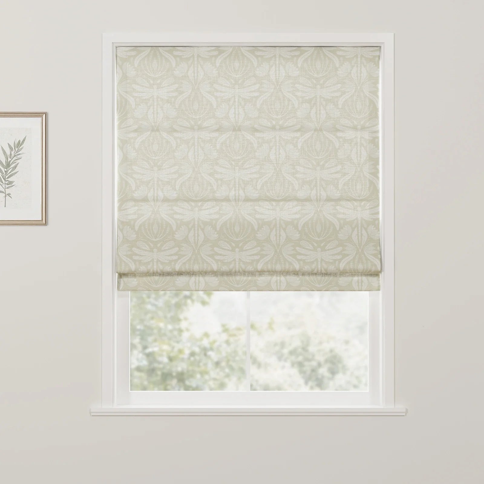 Photograph of a window featuring a cream Roman blind with a prominent stylized dragonfly and foliage pattern, a framed botanical print on the adjacent wall, and a blurred green outdoor scene