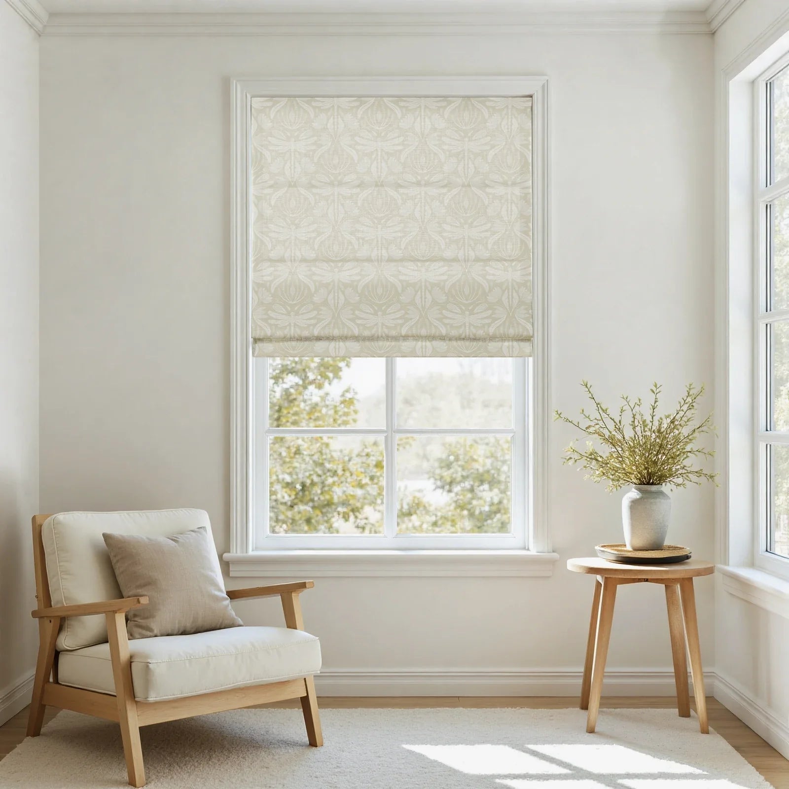 Photograph of a minimalist living space featuring a large window with a cream botanical patterned Roman shade, a natural wood frame armchair with light linen cushions, and a small round side table holding a textured gray vase with fresh green branches