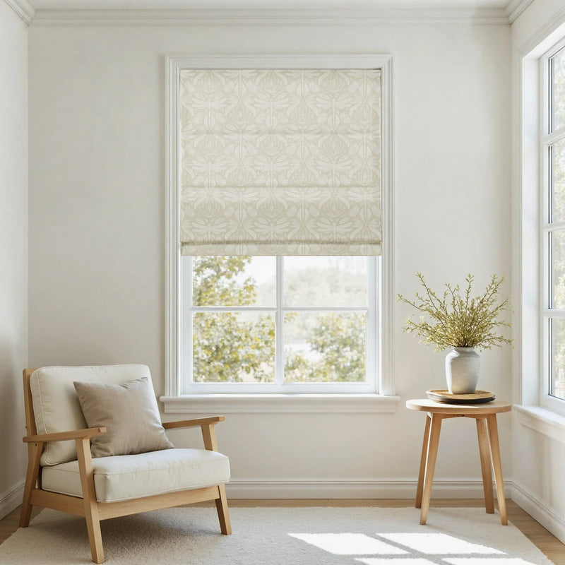 Photograph of a minimalist living space featuring a large window with a cream botanical patterned Roman shade, a natural wood frame armchair with light linen cushions, and a small round side table holding a textured gray vase with fresh green branches