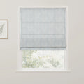 A light blue Roman blind featuring a white leafy damask pattern is partially lowered over a window, revealing blurred green foliage outdoors and a framed botanical print on the adjacent white wall