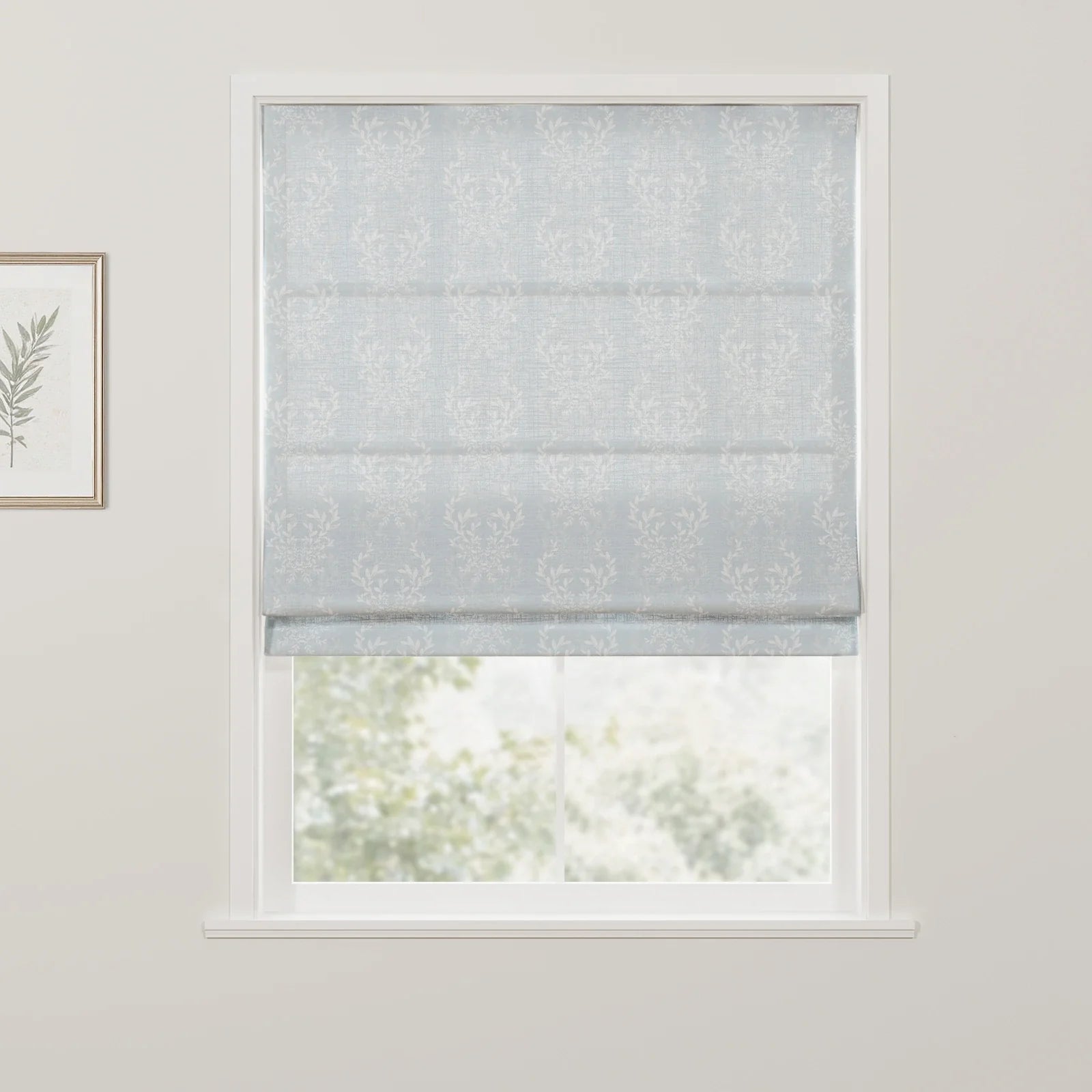 A light blue Roman blind featuring a white leafy damask pattern is partially lowered over a window, revealing blurred green foliage outdoors and a framed botanical print on the adjacent white wall
