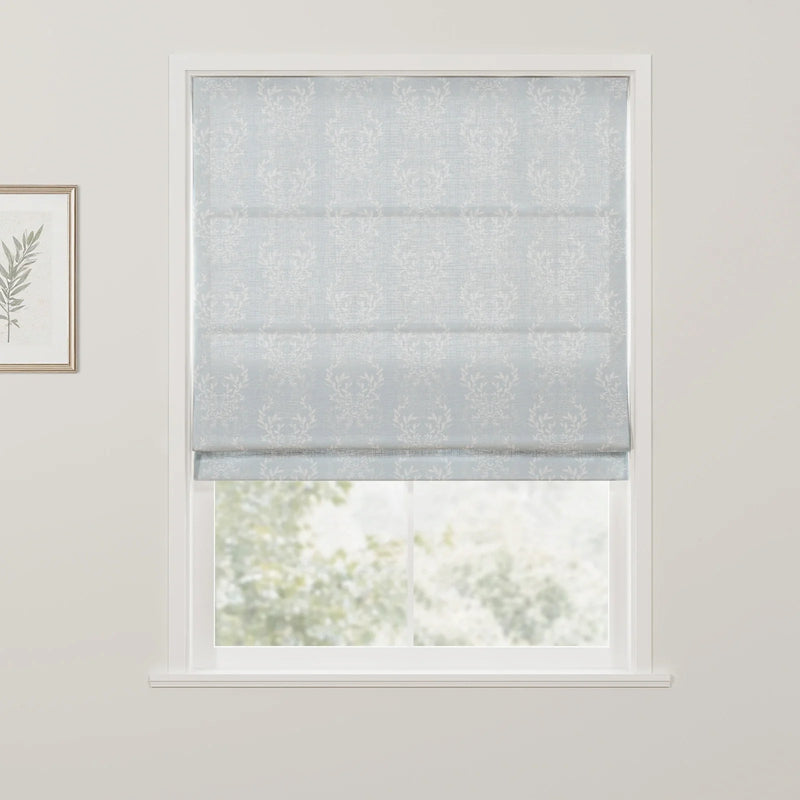 A light blue Roman blind featuring a white leafy damask pattern is partially lowered over a window, revealing blurred green foliage outdoors and a framed botanical print on the adjacent white wall