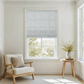 A bright minimalist room featuring a light blue patterned Roman shade over a large window a cream upholstered wooden armchair with a beige pillow and a small wooden side table holding a grey vase with vibrant green foliage